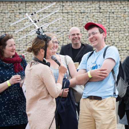 Darton, fully decked out in Radio Local costume including antennae fascinator, interviews a passerby on the street.